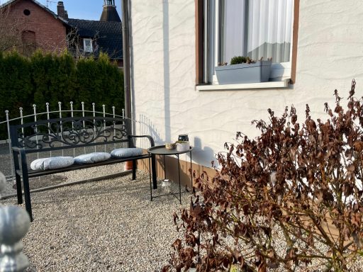 Sitzbank mit Kissen vor einem Fenster, umgeben von einem Garten und einer Hecke.