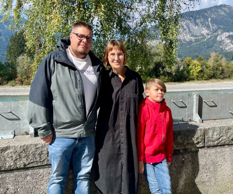 Familie mit Mann, Frau und Sohn am Flussufer des Inn mit Bergen im Hintergrund an einem sonnigen Tag.