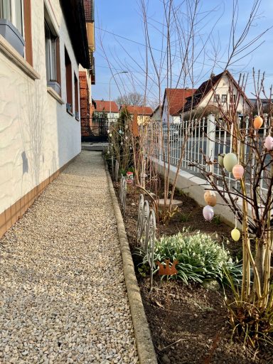 Blick auf einen geschotterten Weg neben einem Garten mit Frühlingsblumen und Ostereiern.
