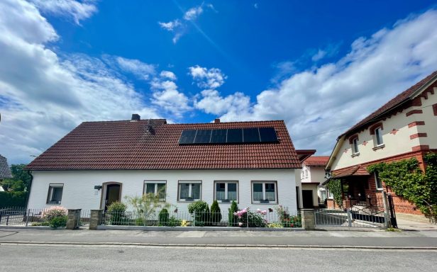 Weißes Haus mit roten Dachziegeln und Solarpanelen, umgeben von blühenden Pflanzen.