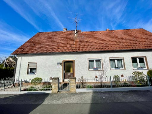Einfamilienhaus mit rotem Schindeldach, braun umrandeten Fenstern, einem gemütlichen Vorgarten und blauer Himmel im Hintergrund.