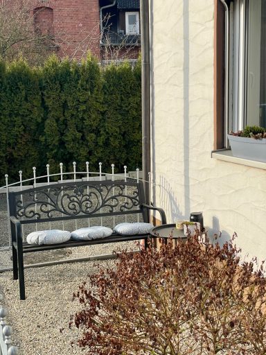 Schwarze Metallbank mit Kissen in einem Garten, umgeben von Pflanzen und einer Mauer.